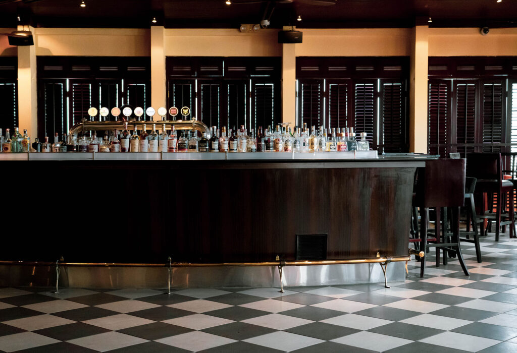 Elegant bar interior with a long wooden counter, rows of liquor bottles, and black-and-white tiled floor.