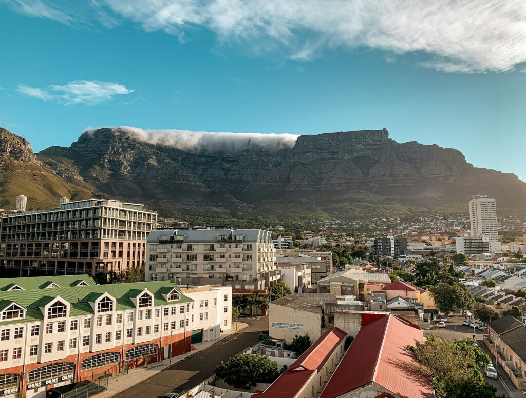 View of Cape Town with Table Mountain in the background and clouds rolling over the top above city buildings.