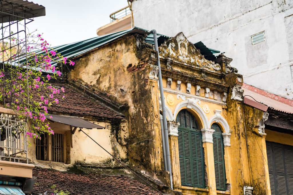 Weathered yellow colonial building with green shutters and bougainvillea flowers in Hanoi, Vietnam.