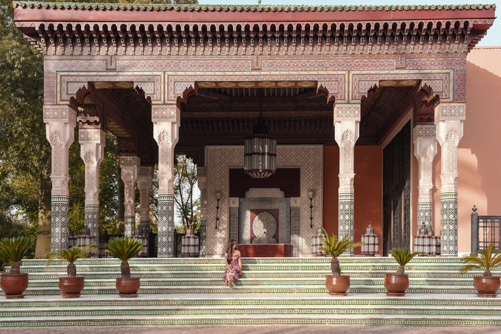 Nastasia Yakoub in a floral dress sitting on zellige-tiled steps of La Mamounia pavilion in Marrakech, surrounded by ornate carved columns and palm trees