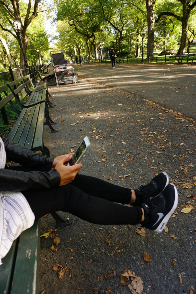 Person sitting on a park bench using a smartphone, wearing black athletic sneakers and leggings in a tree-lined park.