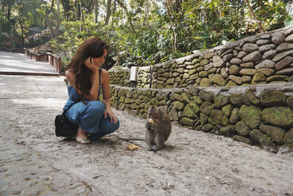 Woman crouching on a forest path watching a monkey eat fruit along a stone wall in a tropical setting.