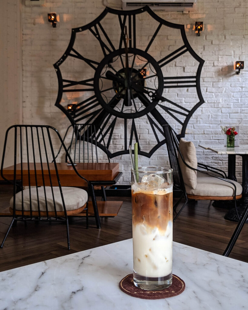 Iced Vietnamese coffee on a marble table inside a café with a large decorative clock on a white brick wall.