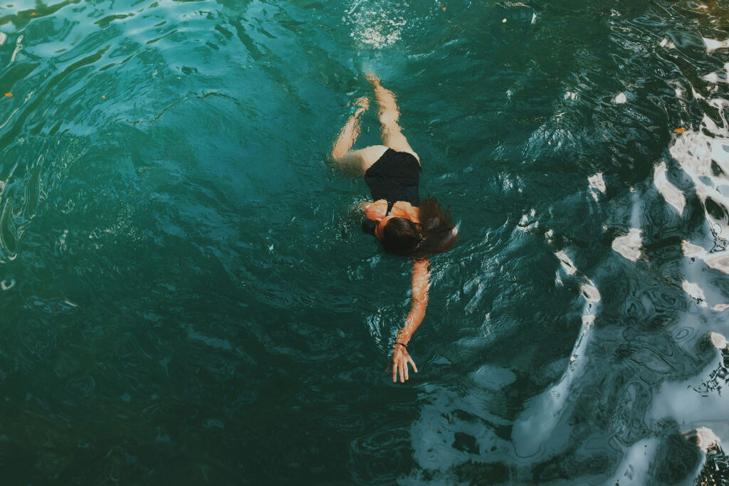 Woman swimming in clear turquoise water, viewed from above, with sunlight reflecting across the surface.