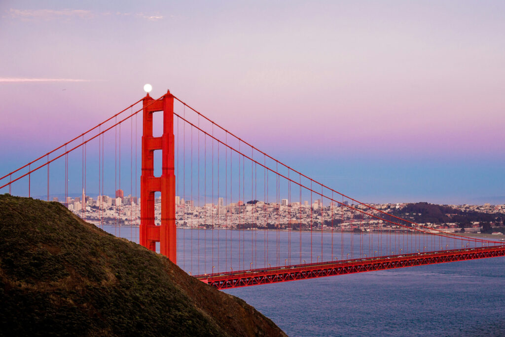 Golden Gate Bridge in San Francisco at dusk with the moon aligned above the tower and city skyline in the background.