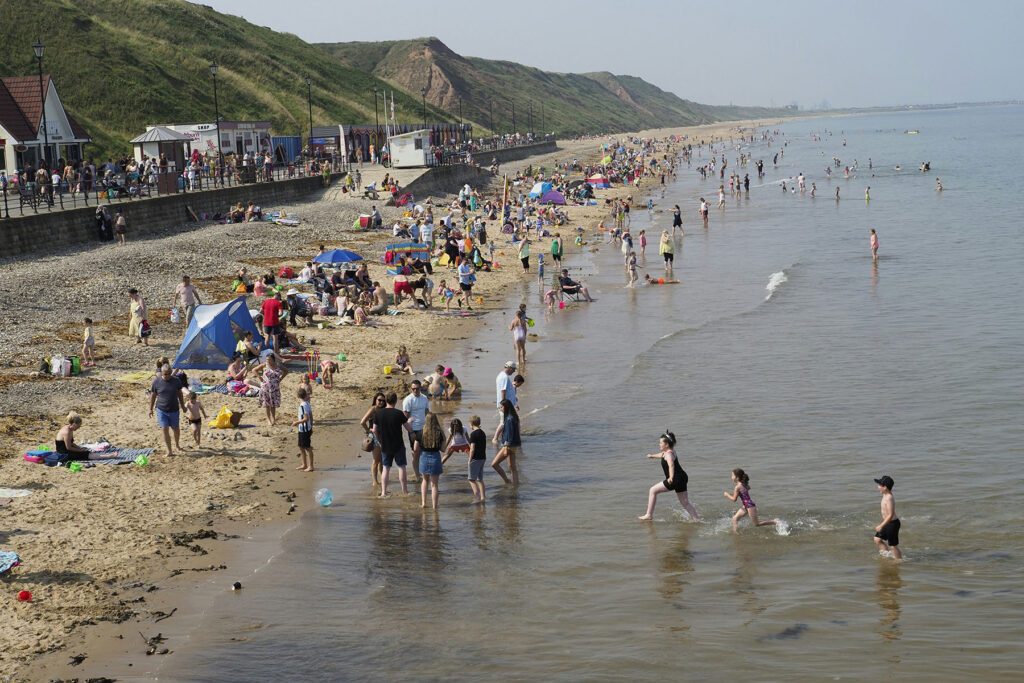 Crowded beach scene with families, children, and swimmers along the shoreline on a sunny day.
