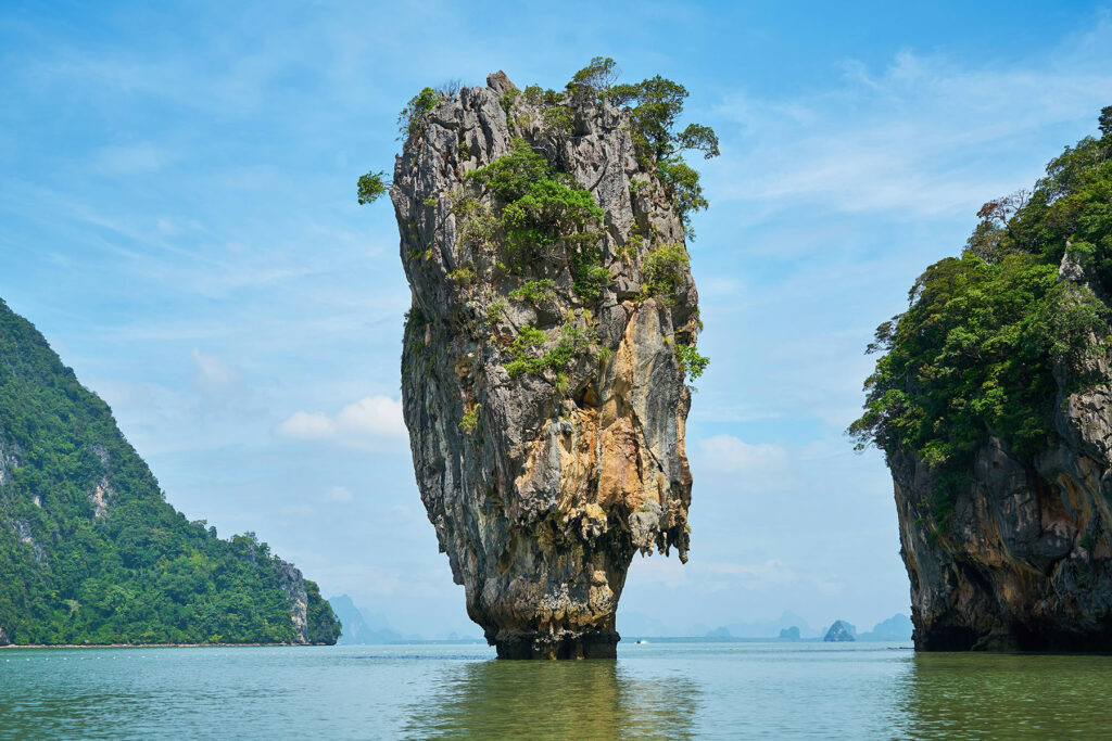 Limestone karst rock formation rising from emerald water in Phang Nga Bay, Thailand, surrounded by lush green cliffs.