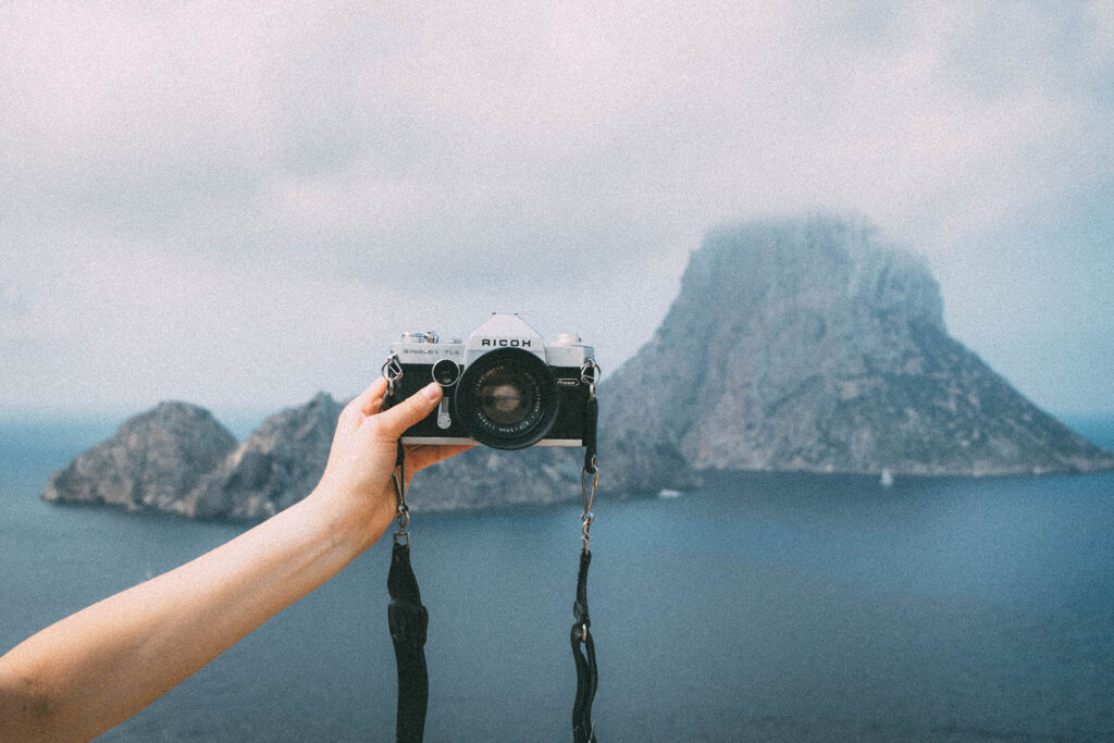 Hand holding a vintage Ricoh film camera in front of a rocky island and calm ocean under a soft, cloudy sky.
