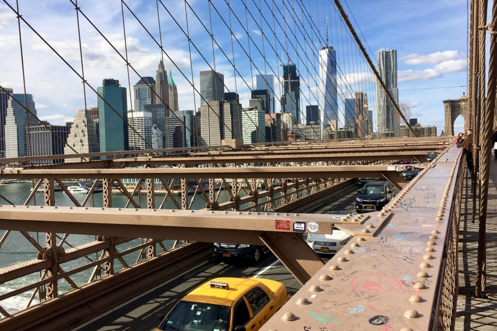 View from the Brooklyn Bridge walkway with cars below and the Lower Manhattan skyline in New York City.