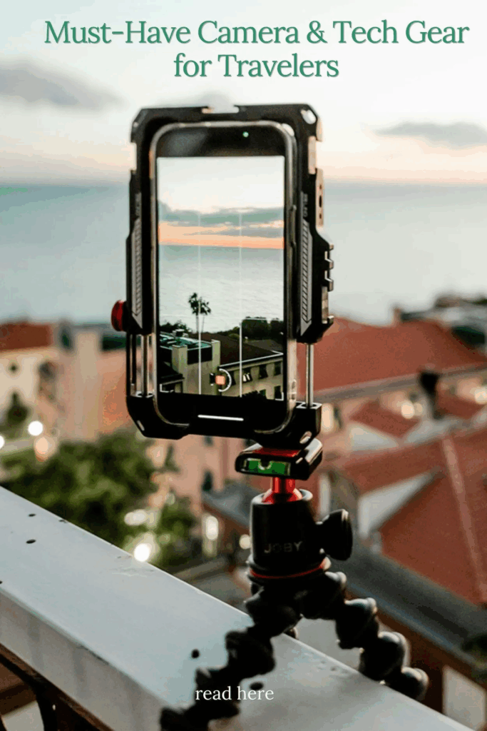 Smartphone on GorillaPod tripod filming Mediterranean sunset over coastal town with terra cotta rooftops, featuring burned-in text overlay reading 'Must-Have Camera & Tech Gear for Travelers' at top and 'read here' at bottom, showcasing best travel tech 2025 for creators