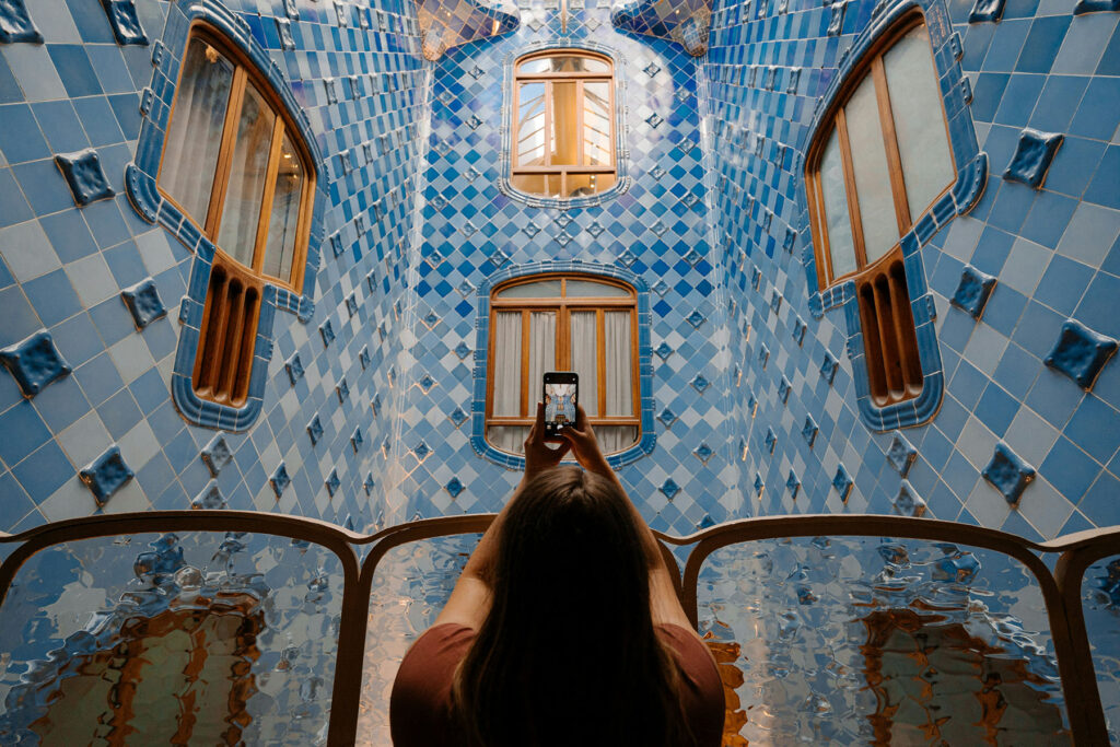 Woman taking a photo inside Casa Batlló in Barcelona with blue mosaic tiled walls and curved windows.