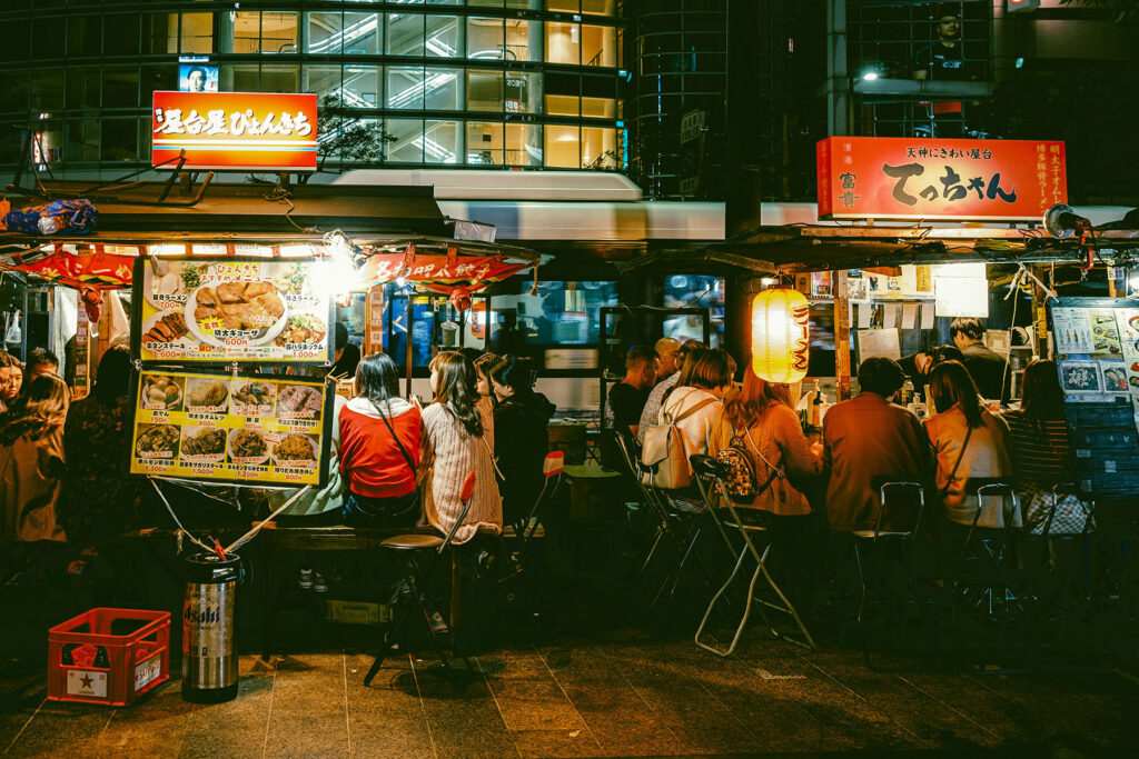 Crowded Japanese street food stalls at night with people seated and eating under glowing lanterns.