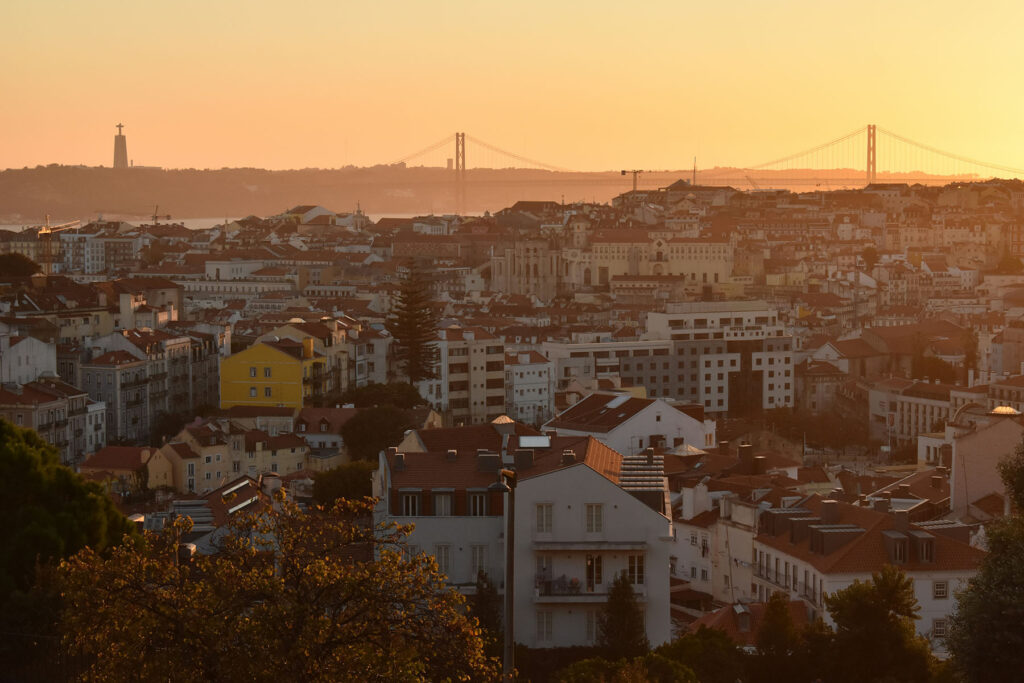 Lisbon cityscape at sunset with terracotta rooftops and the 25 de Abril Bridge in the distance.