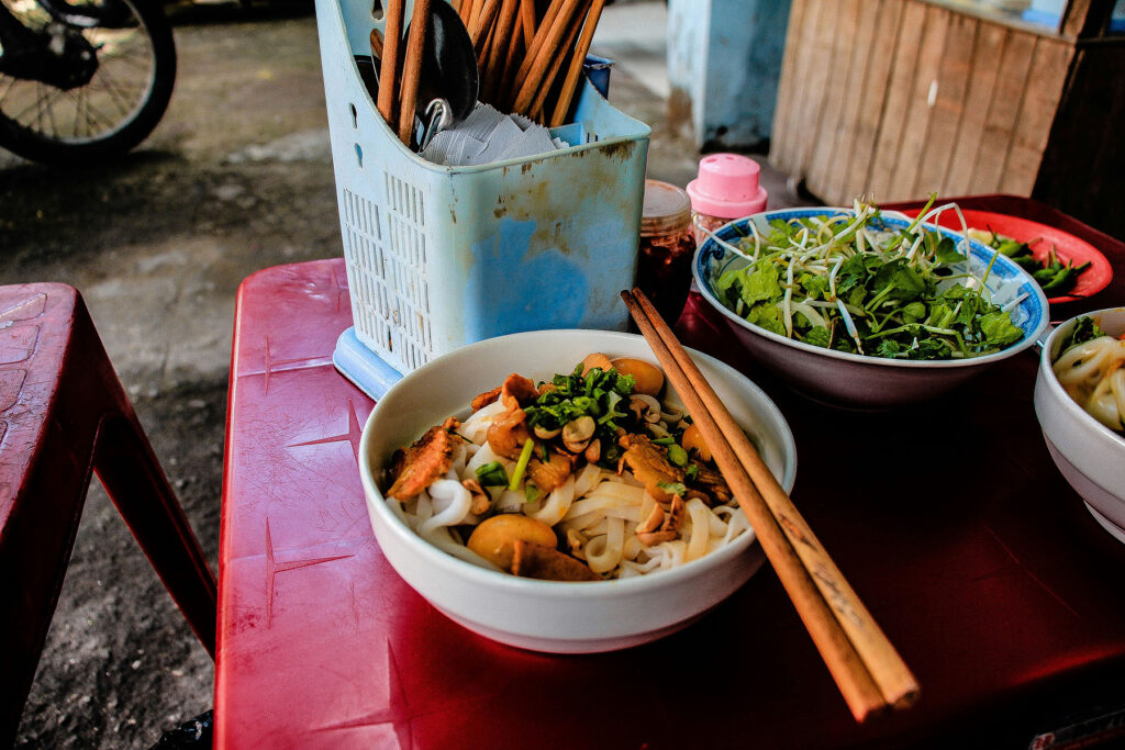Vietnamese noodle dish served on a red plastic table at a street food stall with chopsticks, herbs, and condiments.