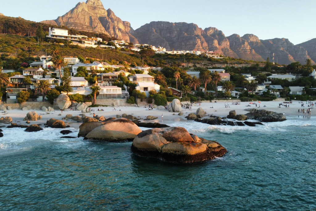 Clifton Beach in Cape Town with large granite boulders, turquoise water, and houses along the hillside at sunset.