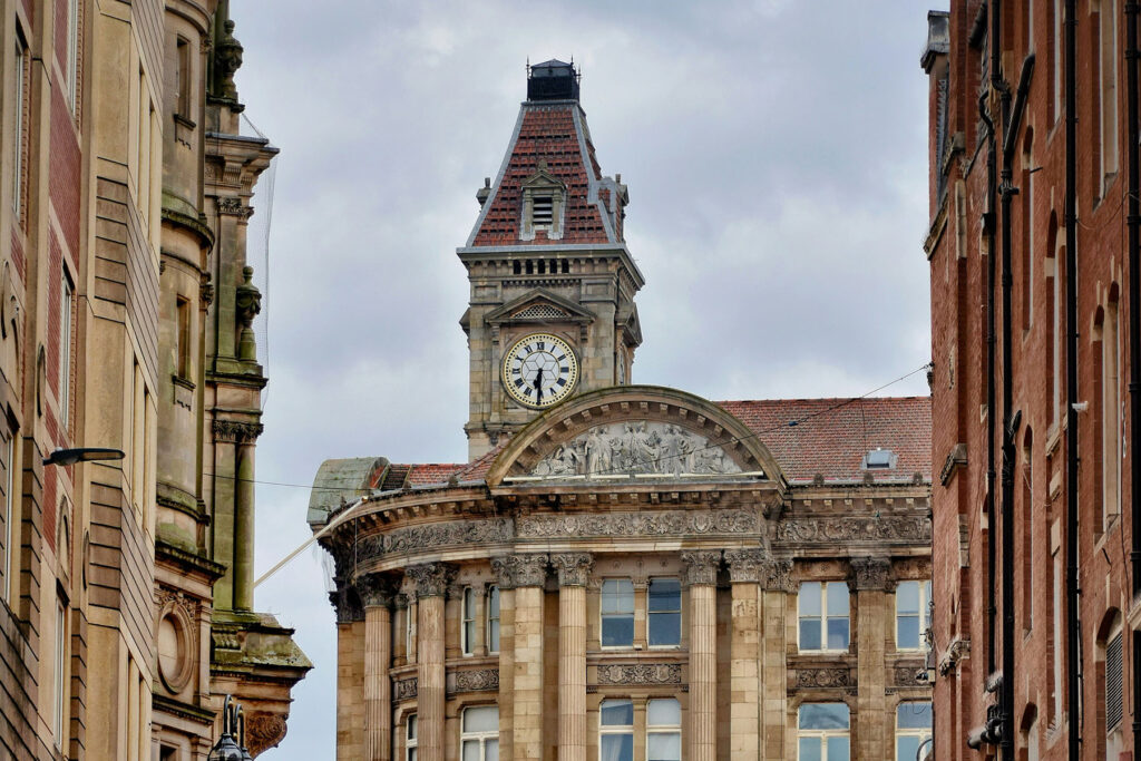 Historic clock tower and ornate building facade in Birmingham city center, framed by surrounding architecture under a cloudy sky.
