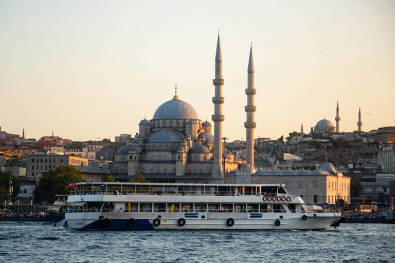 Ferry sailing past a historic mosque in Istanbul at sunset, with domes and minarets glowing in warm golden light along the Bosphorus.