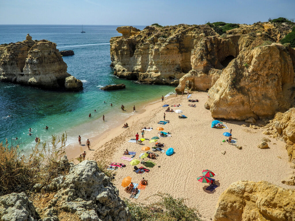 Beach surrounded by golden cliffs in the Algarve, Portugal, with turquoise water and people relaxing on the sand.