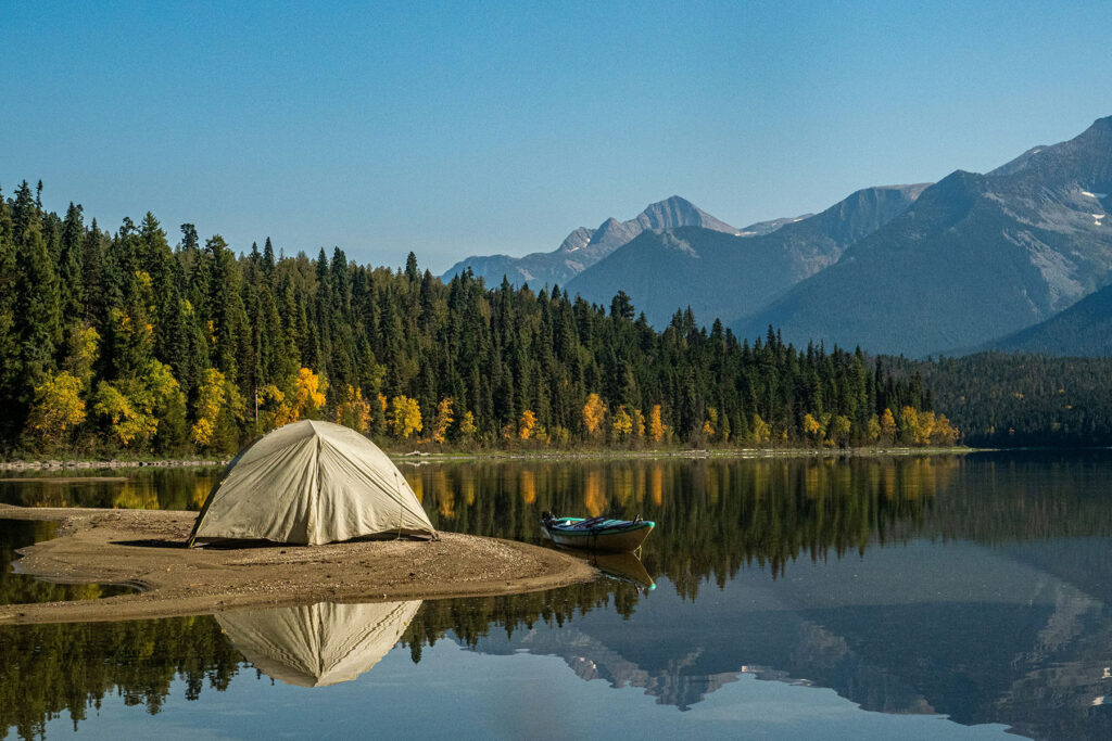 Camping tent by a calm mountain lake with reflections of trees and peaks, surrounded by forest and autumn foliage.
