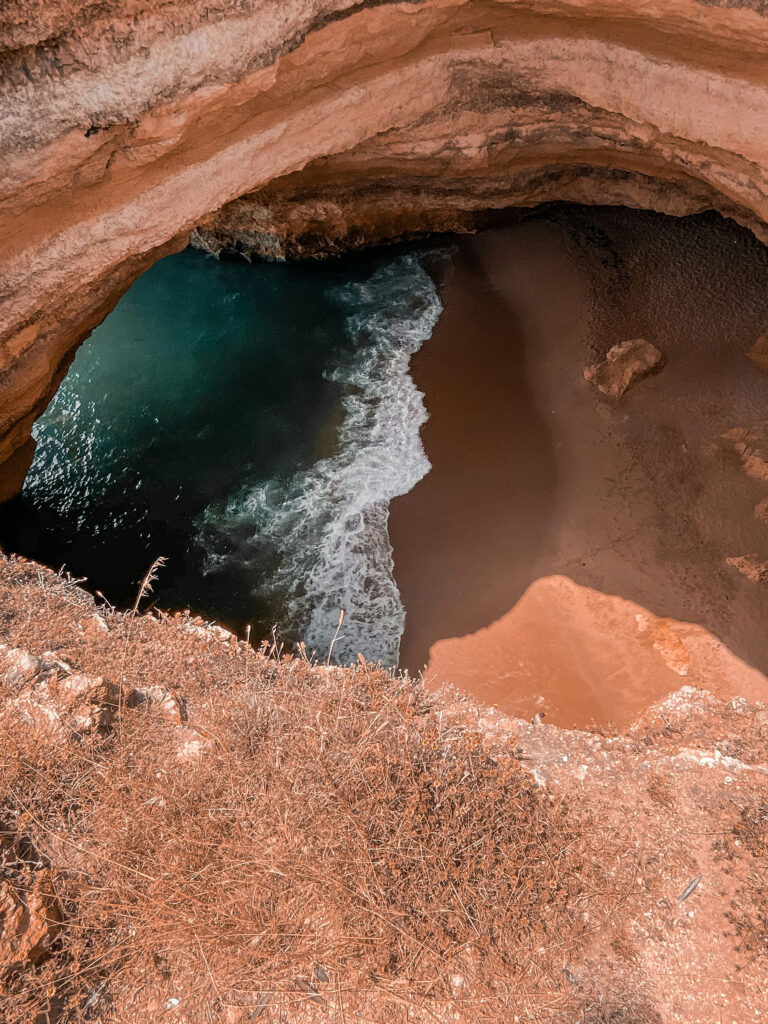 View of a hidden sea cave and secluded beach along the Algarve coast, with waves meeting dark sand beneath layered rock cliffs.
