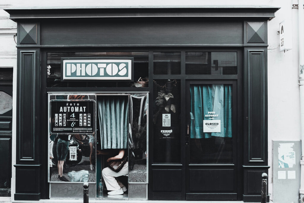 Vintage photo booth storefront with “PHOTOS” sign and curtained booth in a city street setting.