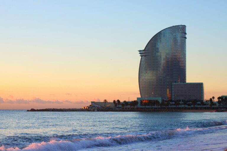 W Barcelona hotel at sunset along Barceloneta Beach, with soft waves in the foreground and pastel sky reflecting on the glass building.