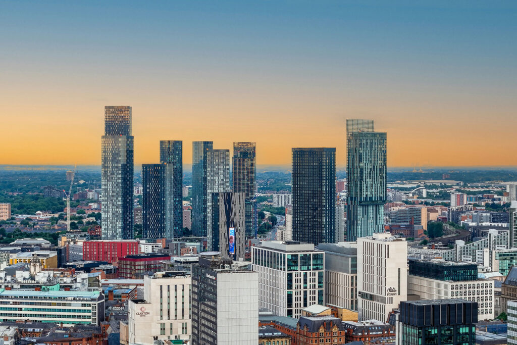Modern skyline of Manchester, England at sunset, featuring tall glass skyscrapers and city buildings under a warm orange sky.
