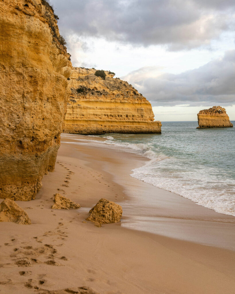 Couple walking along a sandy beach in the Algarve, Portugal, with golden cliffs, ocean waves, and footprints in the sand.