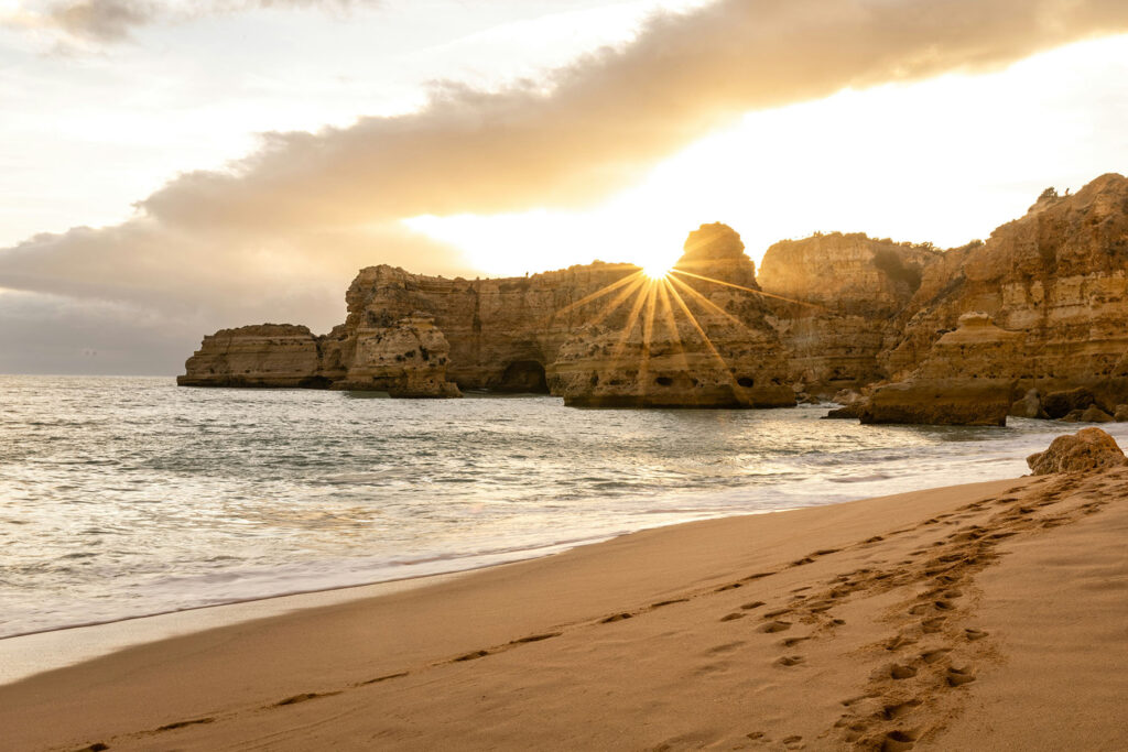 Golden sunset over coastal cliffs and sandy beach in the Algarve, Portugal, with sun rays shining through rock formations.
