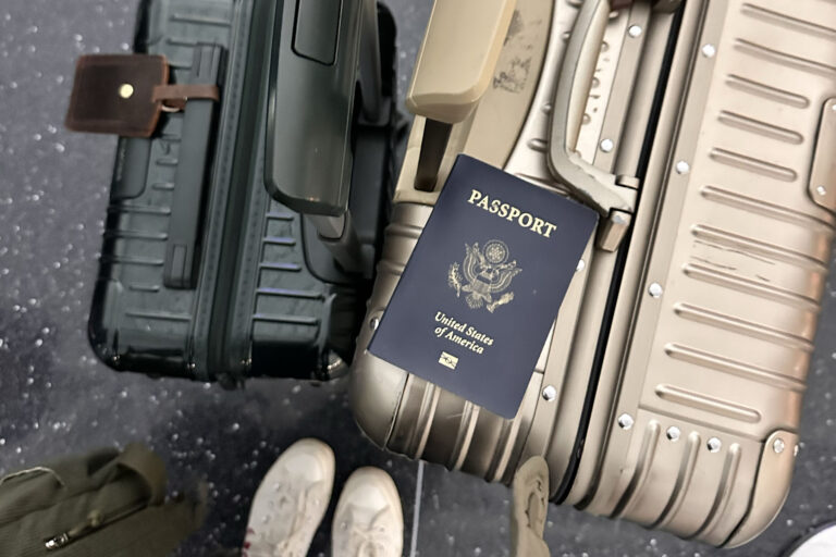 Top-down view of a traveler holding two rolling suitcases—one black and one champagne—with a United States passport resting on the handle, seen against an airport floor with white sneakers visible.