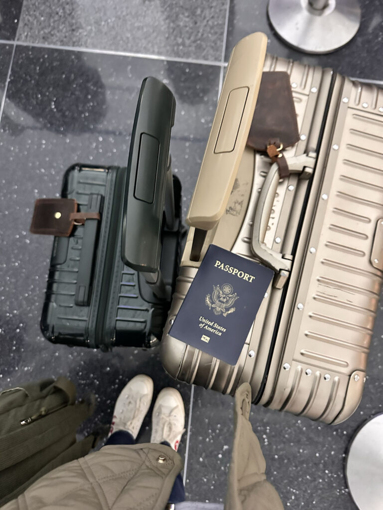 Overhead view of two rolling suitcases one black and one champagne with a U.S. passport resting on the extended handle, traveler’s white sneakers and coat visible on an airport floor.