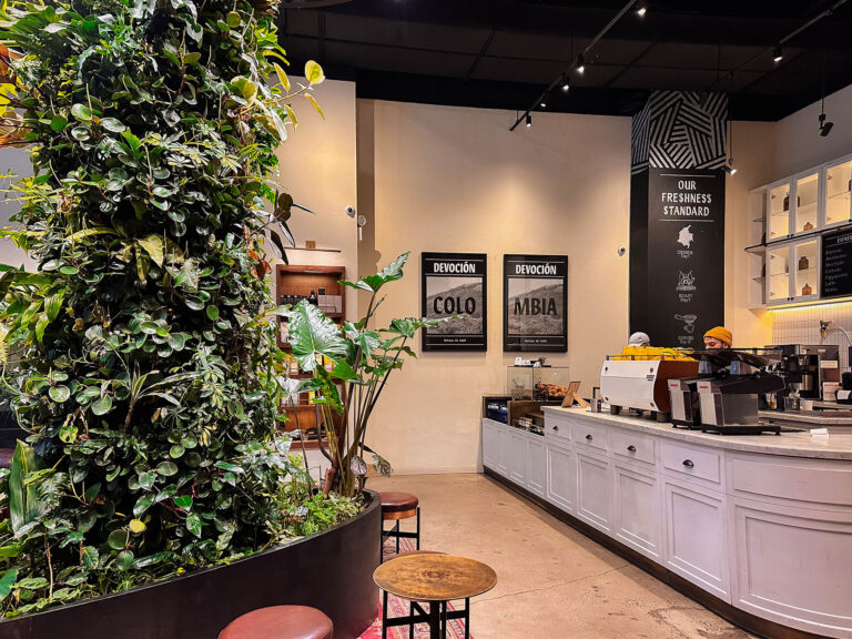 Alt text: Bright, modern interior of a Devoción coffee shop in New York City featuring a lush vertical plant wall, a marble coffee bar with baristas preparing drinks, and minimalist seating. Framed Colombia coffee posters and clean design details create a fresh, botanical atmosphere. Photo by Nastasia Yakoub. All rights reserved. No reposts, reuse, or distribution without written permission.