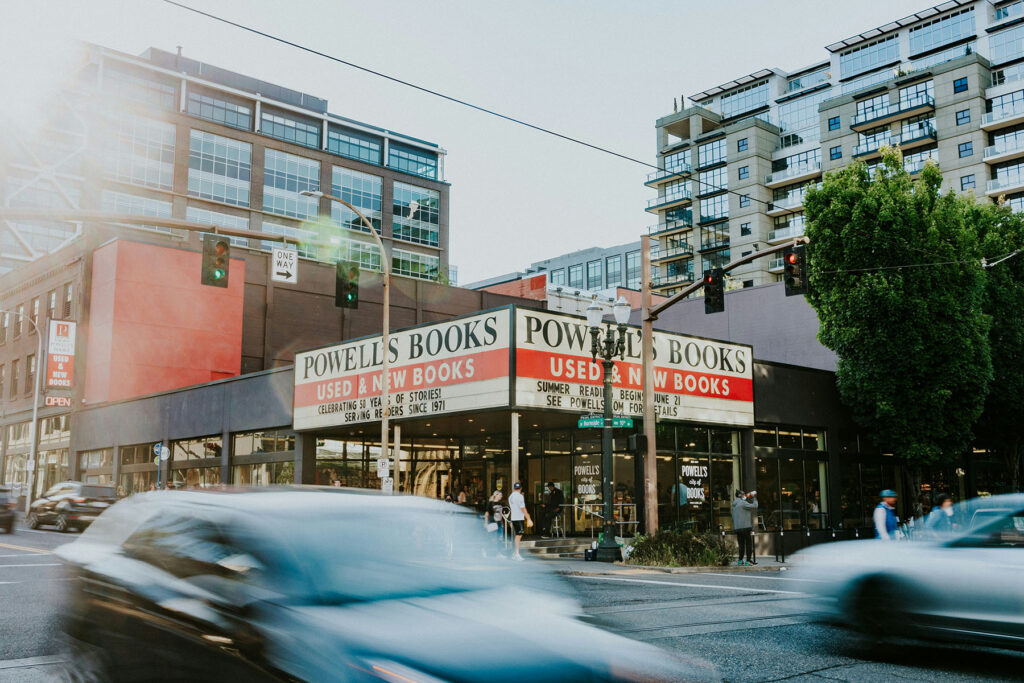 Street view of Powell’s Books in Portland, Oregon, with its iconic “Used & New Books” sign on the corner, pedestrians entering the bookstore, and blurred cars passing by at a busy city intersection in daylight.