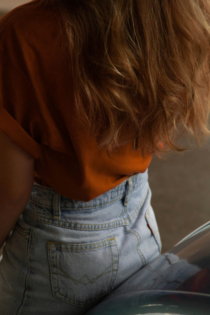**Alt text:**
Close-up of a person with long wavy hair wearing a brown top and light blue jeans, leaning slightly forward in warm, soft lighting.

