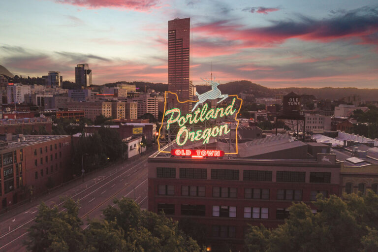 Sunset over downtown Portland, Oregon featuring the iconic “Portland Oregon Old Town” neon sign glowing above historic buildings, with a pink and orange sky and the city skyline in the background.