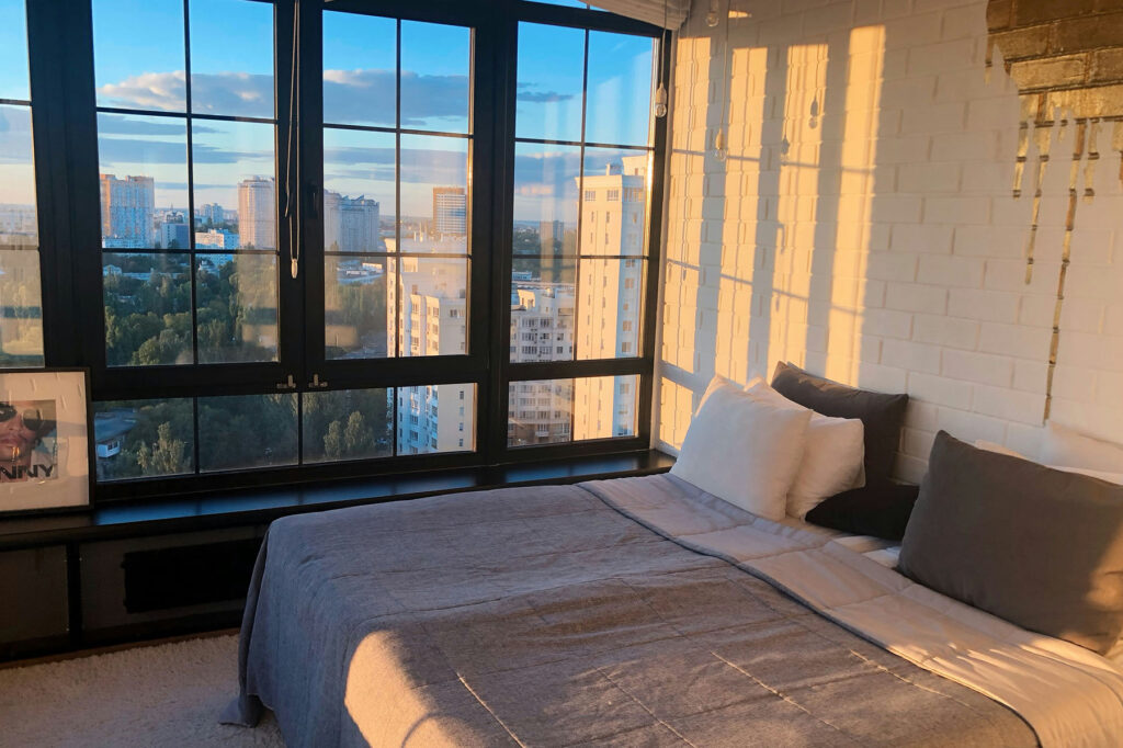 Sunlit bedroom with large windows overlooking a city skyline, featuring a neatly made bed and warm golden hour light.