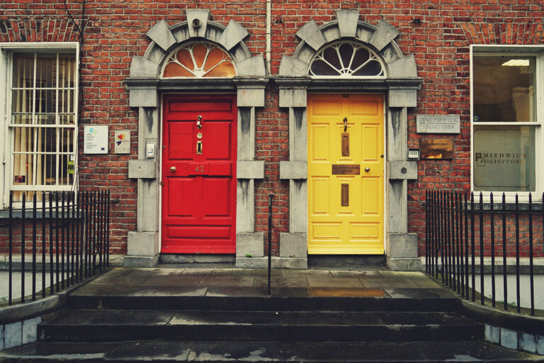 wo colorful front doors one bright red and one yellow—set into a classic red brick building with arched stone frames in Dublin, Ireland. Stone steps lead up to the entrances, with black iron railings and traditional sash windows on either side, creating a charming and symmetrical streetscape.