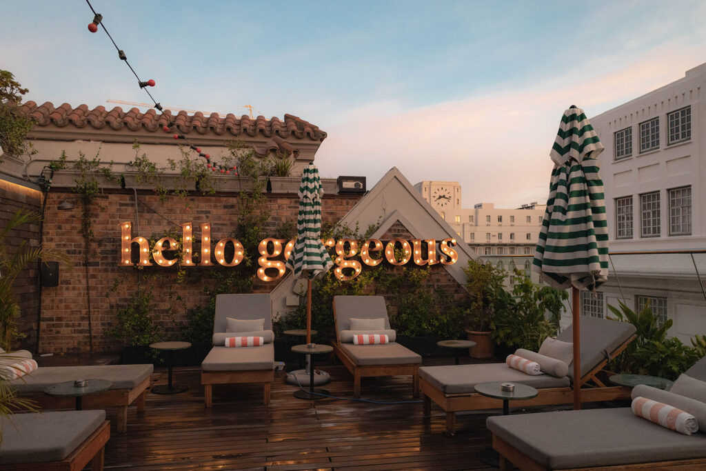 Rooftop pool deck at Gorgeous George in Cape Town featuring striped umbrellas, lounge chairs, glowing marquee signage, and relaxed sunset vibes in the heart of the city. Nastasia Yakoub. All rights reserved. No reposts, reuse, or distribution without written permission.