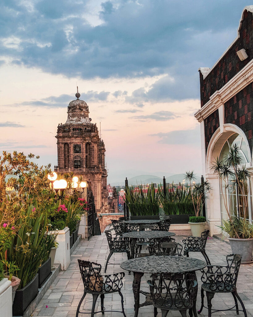 Rooftop terrace at Hotel Zócalo Central in Mexico City overlooking the historic Metropolitan Cathedral at sunset, with wrought iron tables, lush greenery, and dramatic evening skies. Nastasia Yakoub. All rights reserved. No reposts, reuse, or distribution without written permission.