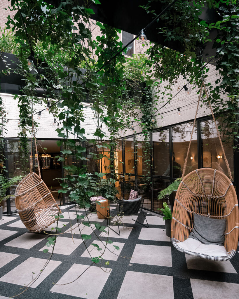 Lush indoor courtyard at Kimpton De Witt Amsterdam featuring hanging greenery, woven swing chairs, black-and-white tiled flooring, and a cozy boutique hotel atmosphere. Nastasia Yakoub. All rights reserved. No reposts, reuse, or distribution without written permission.