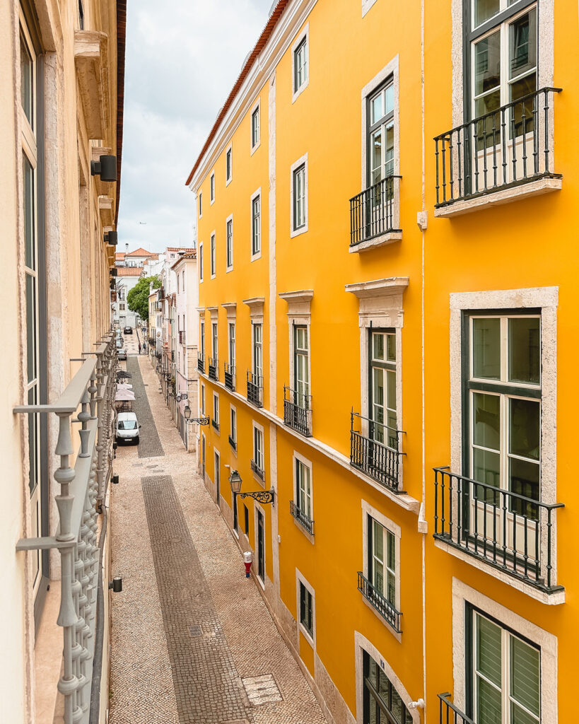 Narrow cobblestone street in Lisbon lined with vibrant yellow buildings, wrought-iron balconies, and classic Portuguese architecture viewed from above. Nastasia Yakoub. All rights reserved. No reposts, reuse, or distribution without written permission.