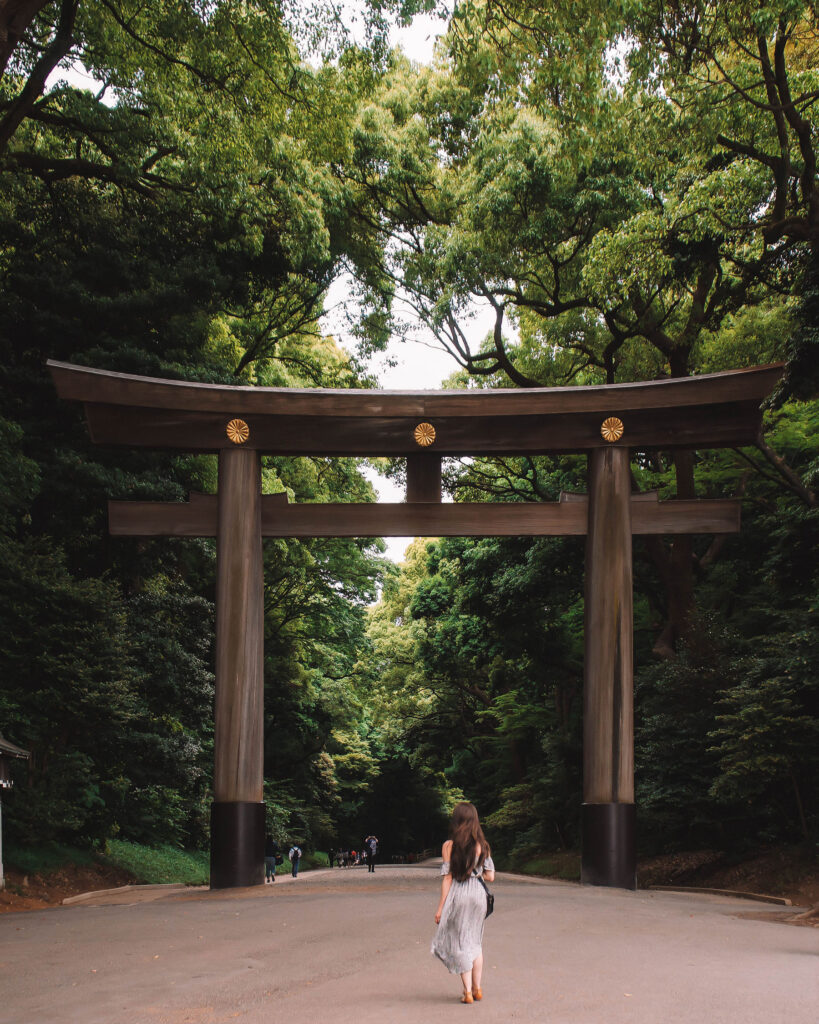 Woman walking beneath the towering wooden torii gate at Meiji Shrine in Tokyo, surrounded by lush green forest and peaceful pathways. Nastasia Yakoub. All rights reserved. No reposts, reuse, or distribution without written permission.
