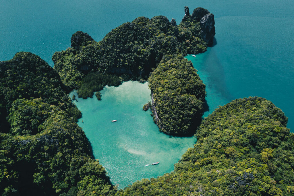 Aerial view of lush tropical islands surrounded by crystal-clear turquoise water with small boats drifting through a secluded lagoon in Thailand.